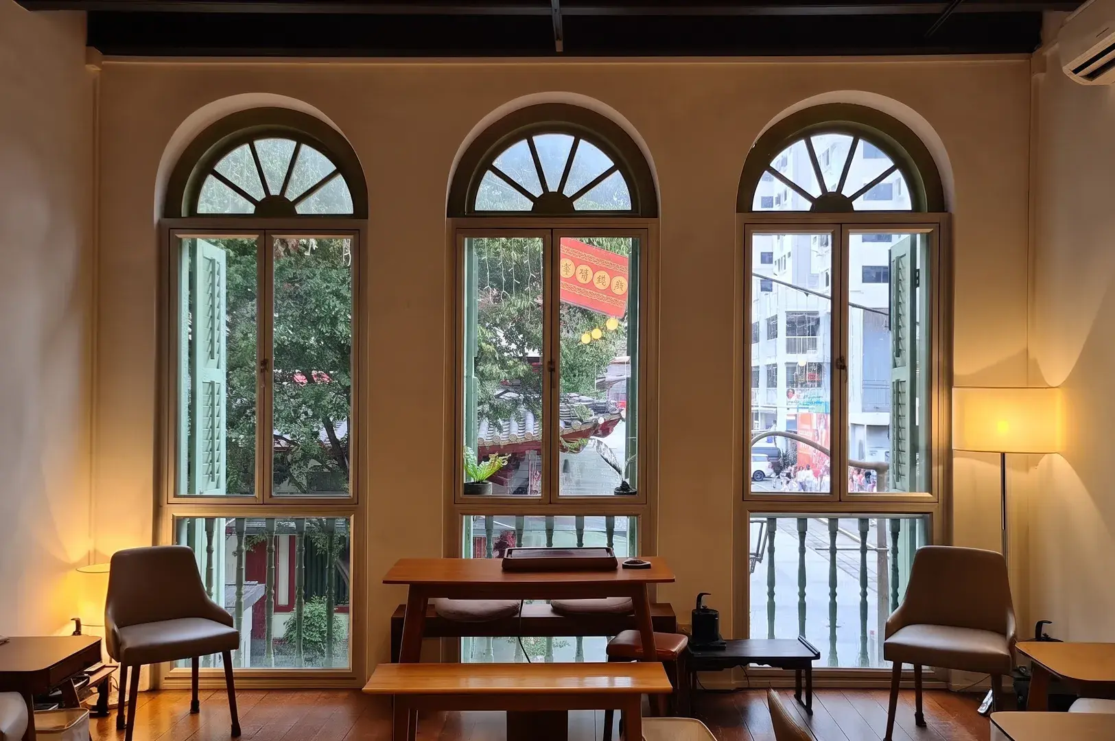 Three arched shophouse windows overlooking city streets from a minimalist room with wooden furniture.