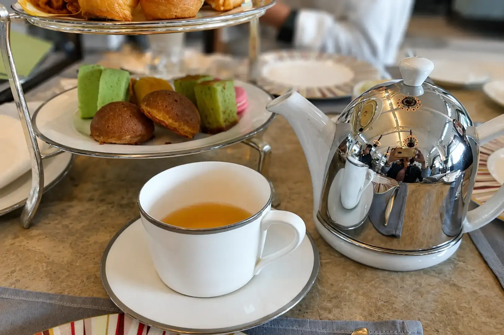 A close-up view of a teacup filled with light amber tea sitting on its white saucer next to a highly reflective, polished chrome teapot, with a partially visible silver tier stand filled with colorful pastries in the background.