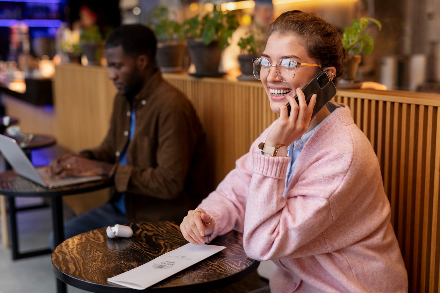 A smiling woman in a pink sweater sits in a café, talking on her smartphone while enjoying a relaxed atmosphere. This illustrates how modern contact center solutions enable remote omnichannel customer support.
