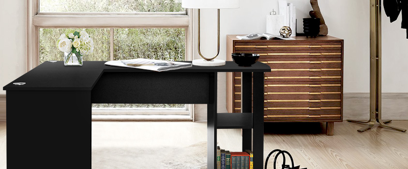An Artiss Black Corner Student Office Desk set in a brightly lit room. The desk is holding a magazine, white table lamp, some books and a glass vase with white roses. In the background is a wood and gold buffet sideboard and a brass coat hanger.