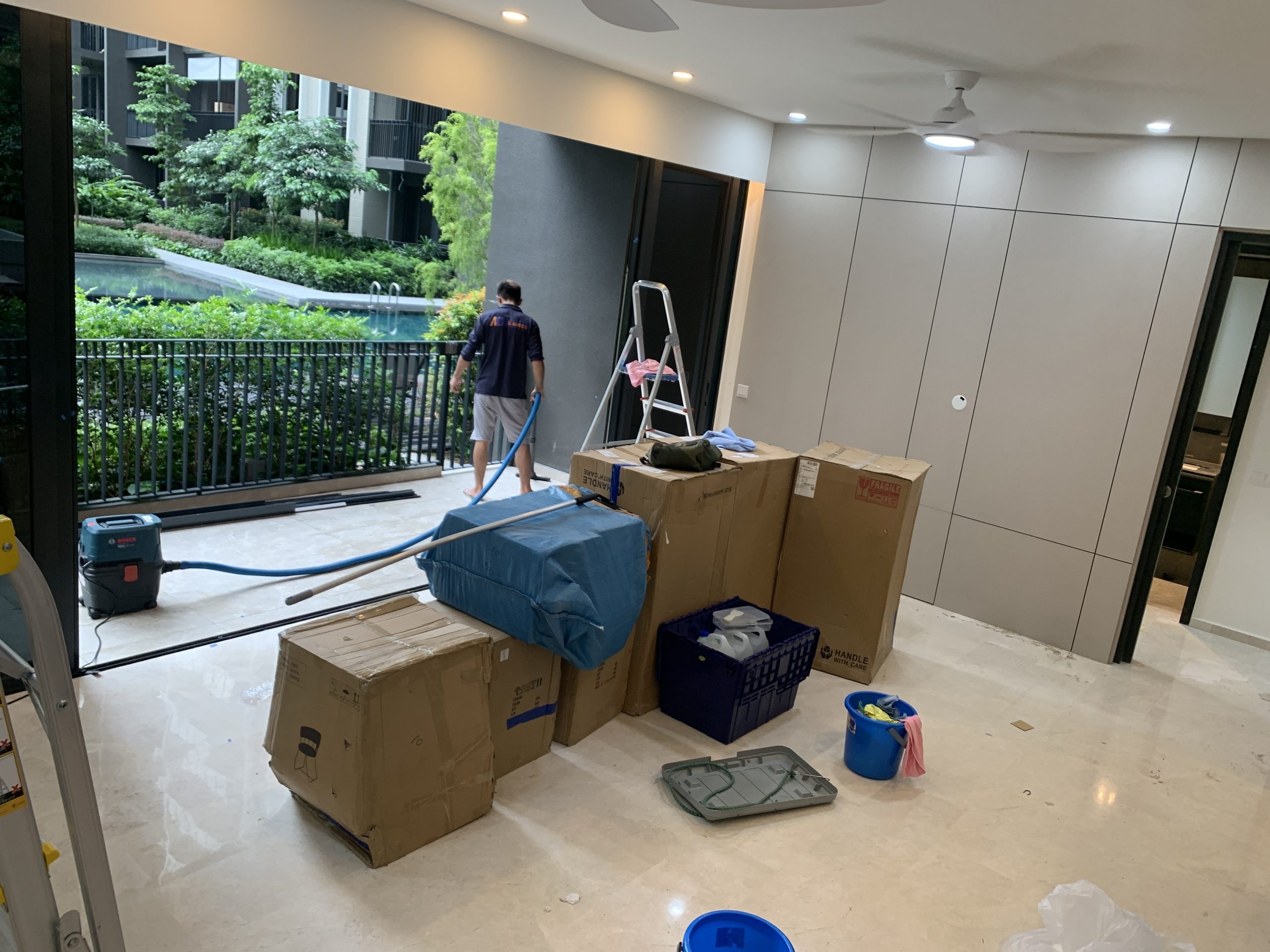 Interior of a modern apartment with several packed cardboard boxes, cleaning supplies, and a vacuum hose on the floor; a person stands outside near a small pool or garden area, likely preparing for a cleaning or moving task