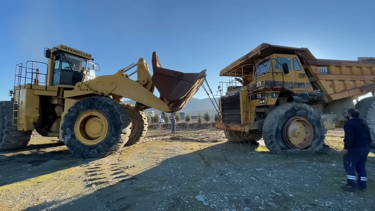 A Caterpillar wheel loader lifting the bucket of another Caterpillar wheel loader while a person watches on at a construction site