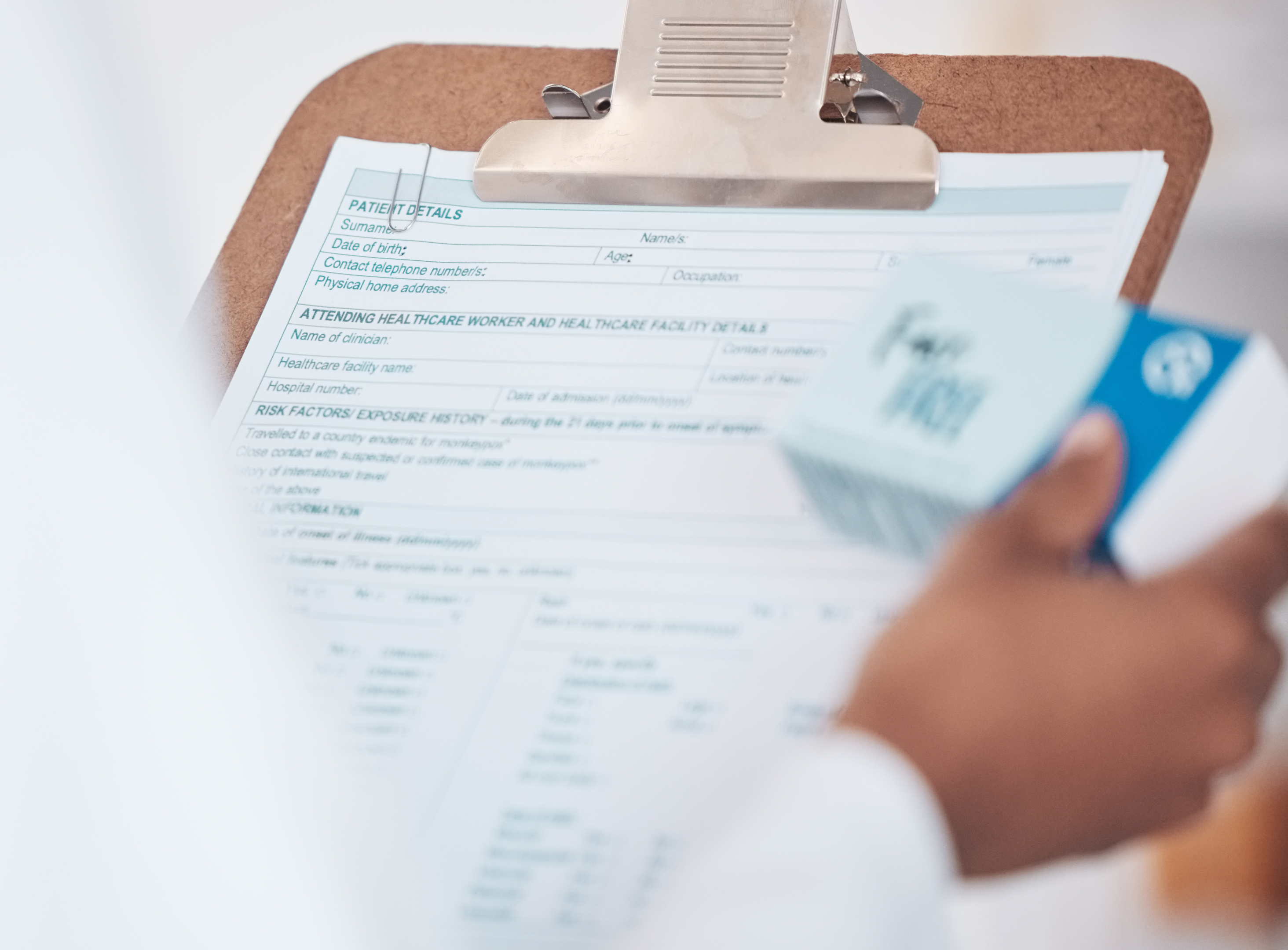 A closeup of a patients hand holding a Medical record form on a clip board and a box of medicine in the other hand. 