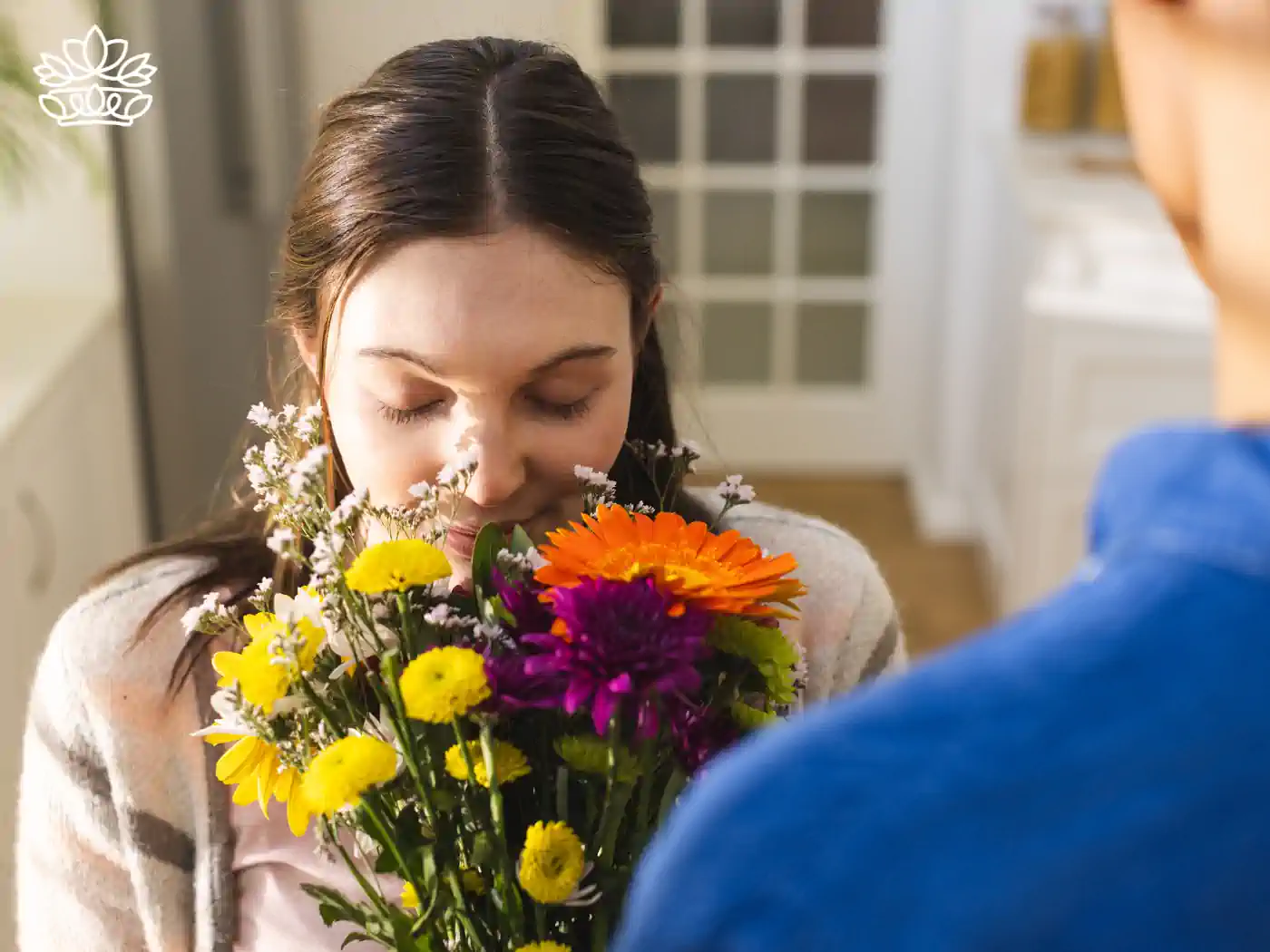A woman joyfully smelling a colorful bouquet of fresh flowers including orange gerberas and yellow daisies, gifted by a person visible only from the back.