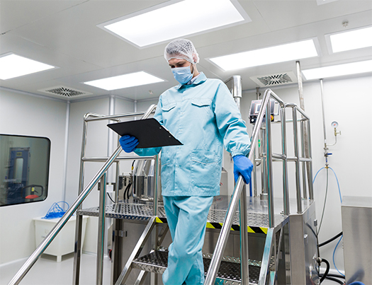 A technician in a cleanroom suit, mask, and gloves holding a clipboard while descending stairs in a manufacturing facility.