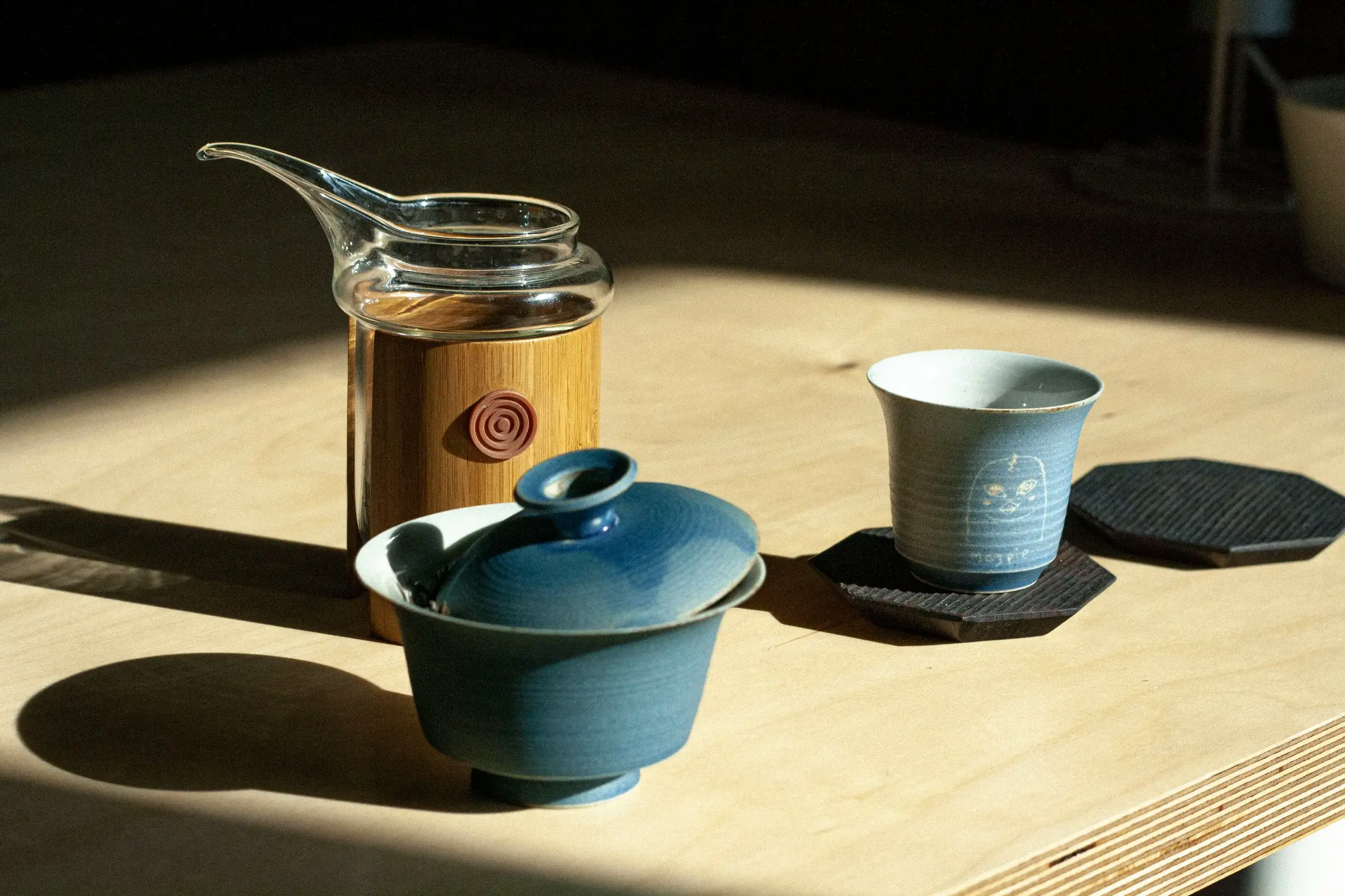 A blue ceramic gaiwan and a matching teacup sit on a light wooden table, illuminated by sharp afternoon sunlight. A glass pitcher with a bamboo sleeve stands behind them, casting a long shadow across the surface.