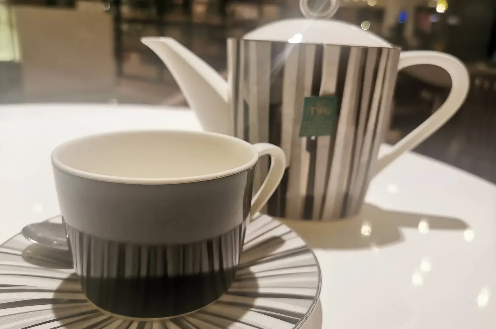 A stylish, black and white striped porcelain teapot and matching teacup are set on a pristine white table, reflecting the elegant theme of the high tea experience. The teapot has a small green tag, and the teacup sits on a matching saucer, emphasizing the sophisticated and coordinated design of the tea ware against the clean, minimalist background.