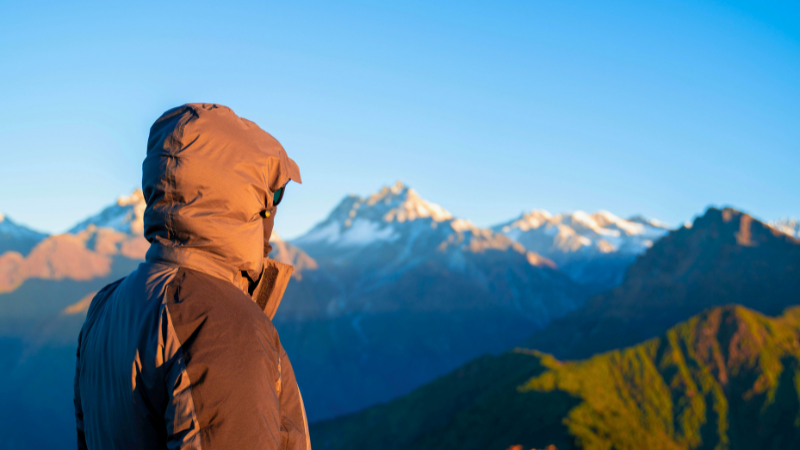A hooded gorpcore jacket wearer gazing at a vast mountain range.