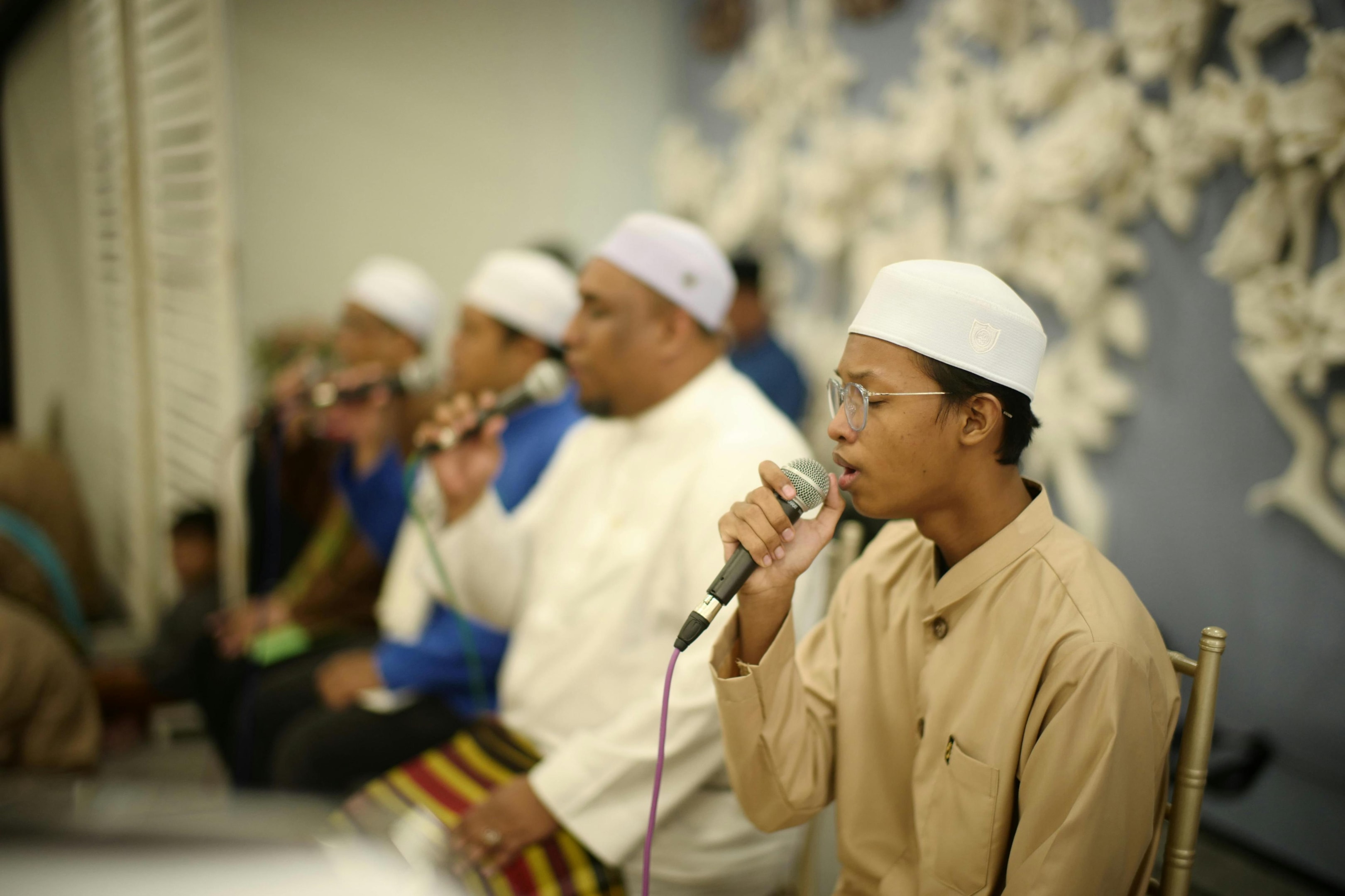 A group of men wearing traditional tunics and white skullcaps sit in a row, each holding a microphone as they perform together. The camera focuses on a young man in the foreground dressed in beige who is singing with expression, while the others accompany him against a decorated wall background.