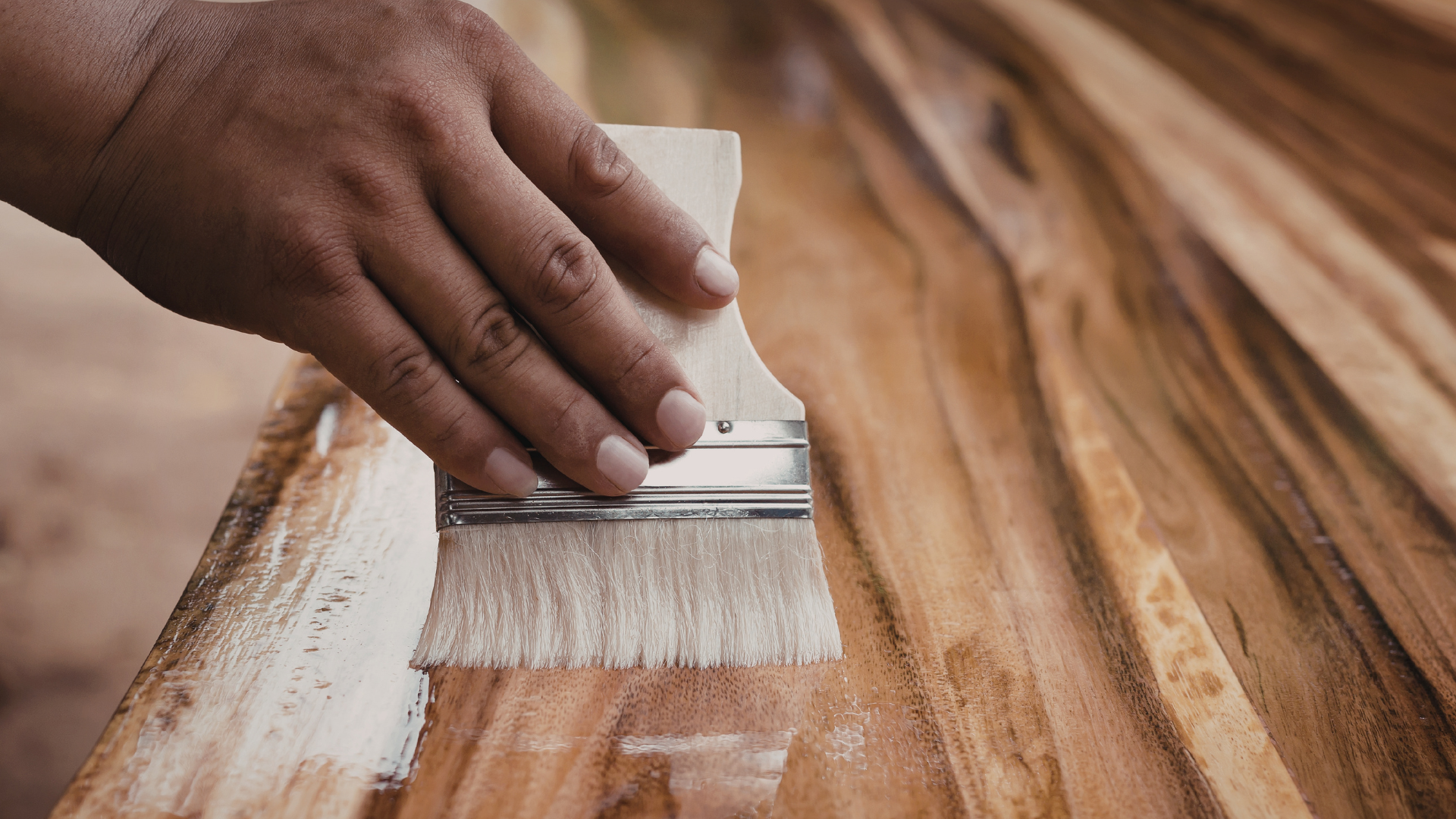 A hand applying a layer of varnish on a piece of wood using a paint brush.