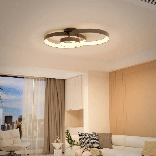 Modern living room with a circular ceiling light fixture featuring overlapping LED rings, illuminating a cozy space with a white sectional sofa, beige accent wall, and large window with sheer curtains.