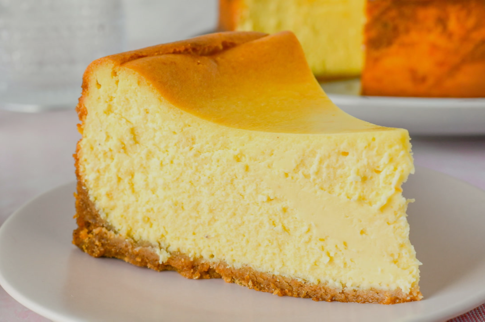 A creamy, golden slice of cheesecake with a thick crust sits on a white plate. The cake's rich texture and color convey a sense of indulgence.
