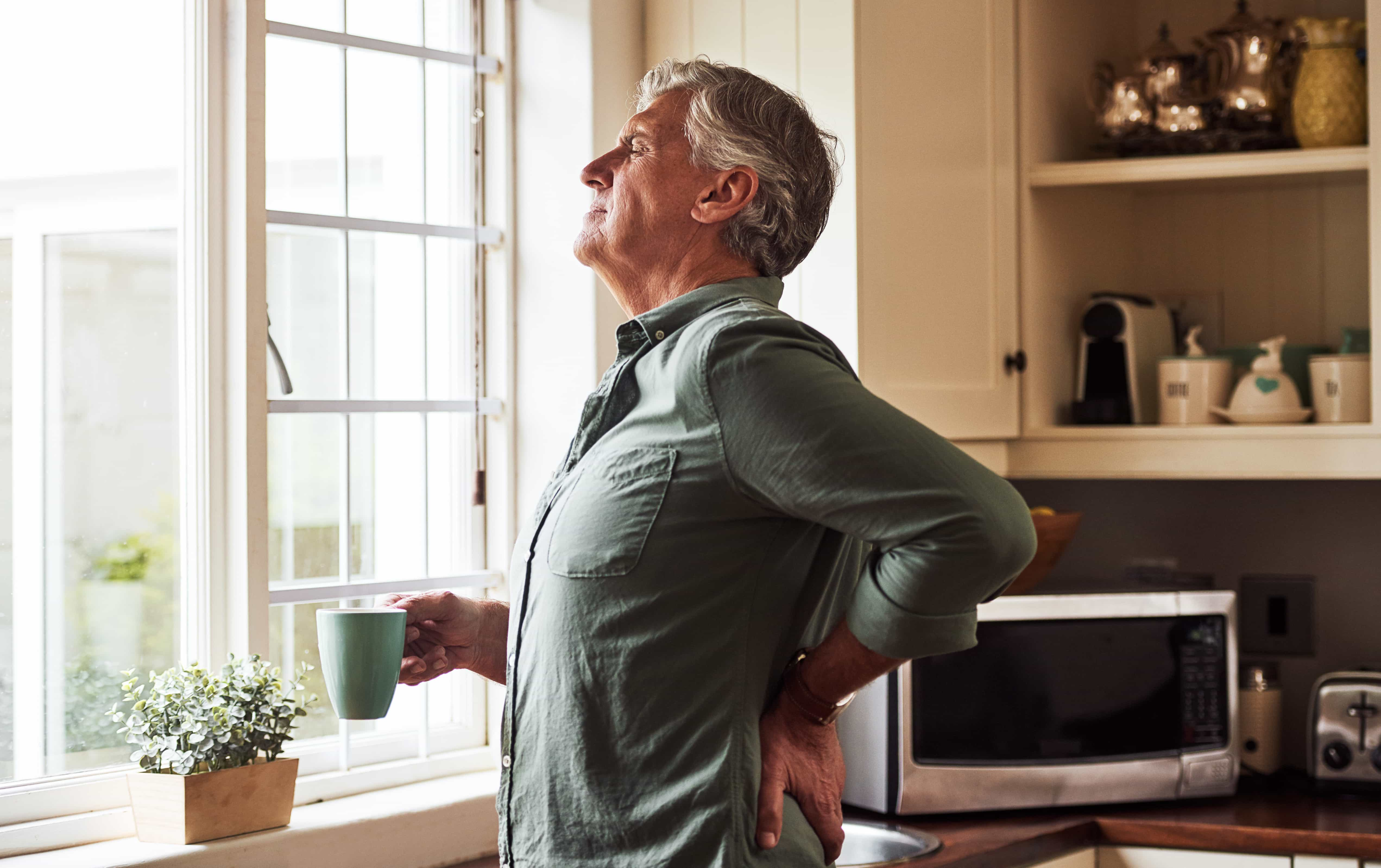 Older man experiencing chronic aching and stiffness in the morning. He is holding a coffee and clutching his lower back in pain.