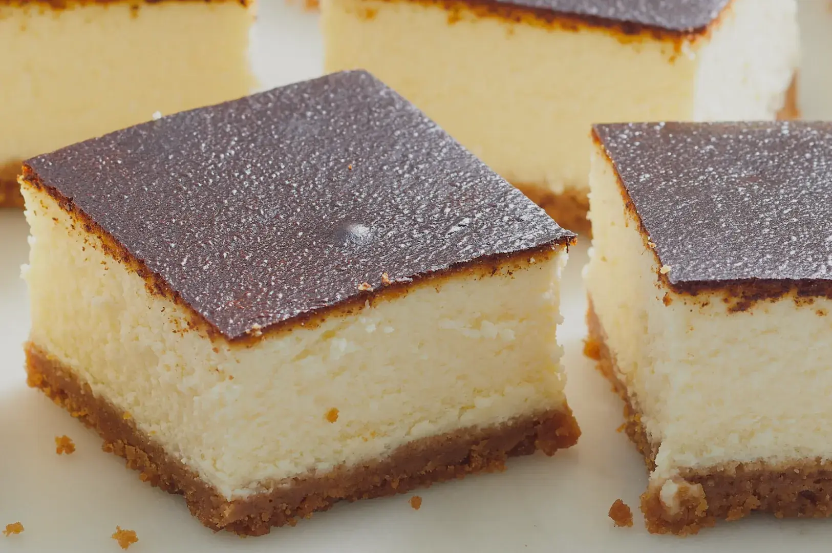 A close-up of square slices of cheesecake with a chocolate ganache topping and a crumbly crust.