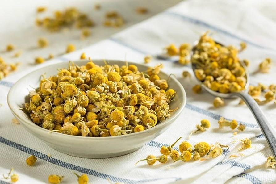 This image shows a small ceramic bowl filled with dried chamomile flowers, set against a light-colored cloth with blue stripes. Scattered blossoms and a metal spoon resting nearby complete the bright, rustic kitchen scene.