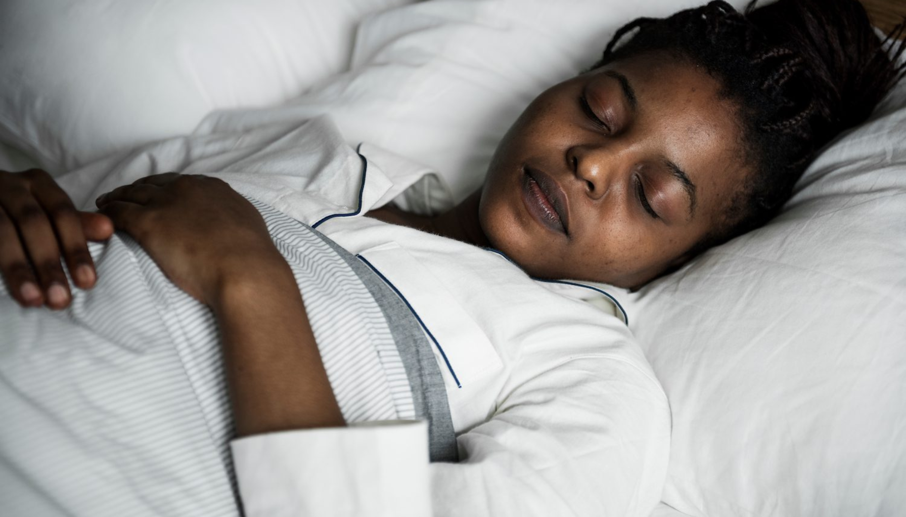 Kvinne med flettefrisyre sover rolig i seng med hvite laken, iført hvit pyjamas med mørk kant.