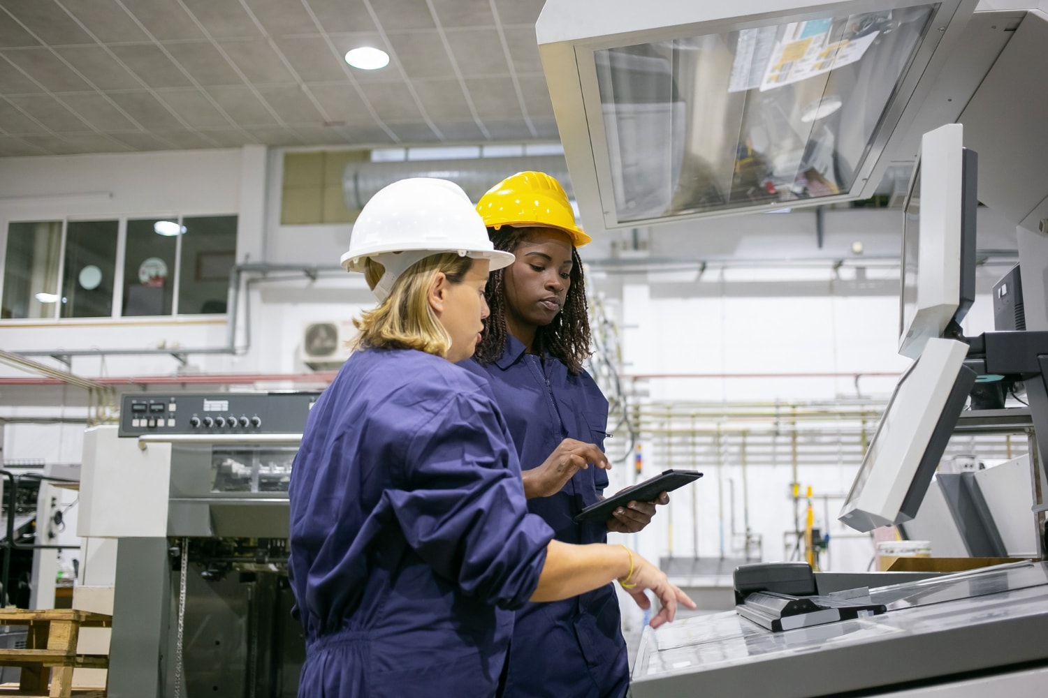 Two female workers in hard hats and blue coveralls consult a touchscreen interface on a large printing machine. Unified Communications powered by UCaaS enables remote system control and diagnostics through cloud telephony and real-time cloud-based communication, enhancing operational oversight.