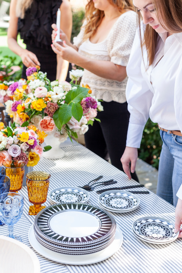 Women arranging patterned plates and glassware on a long table decorated with fresh flowers, preparing a styled tablescape for a gathering.