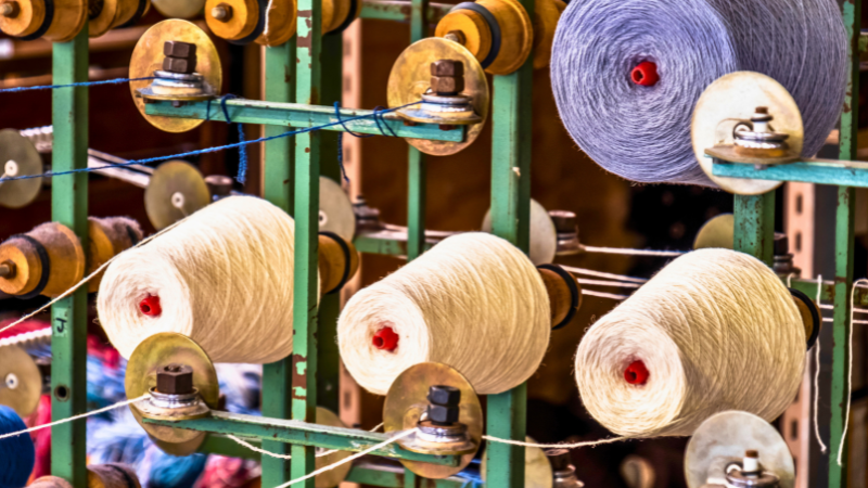 A close-up of spools of polyester yarn on a machine, with cream-colored and grey yarn visible.