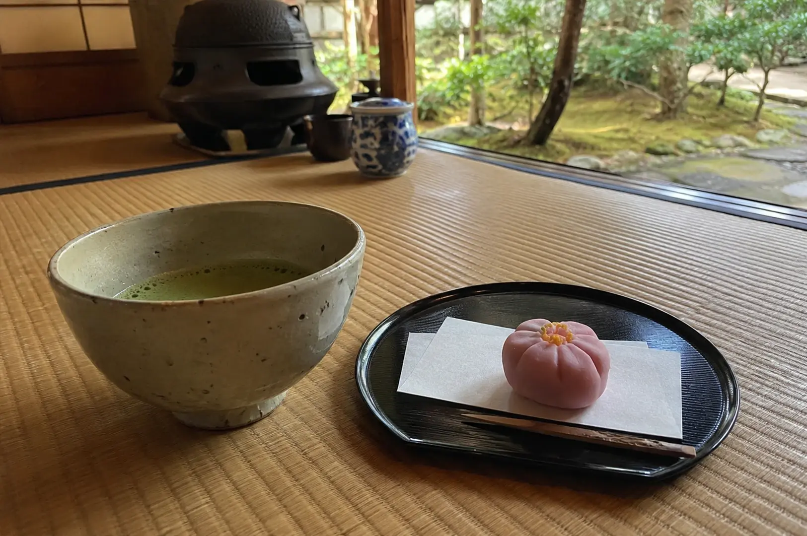 The arrangement is set on tatami mats within a traditional Japanese room, with a soft-focus background of a peaceful garden visible through an open doorway. The lighting is natural and gentle, highlighting the texture of the matcha foam and the precise craftsmanship of the seasonal confection.