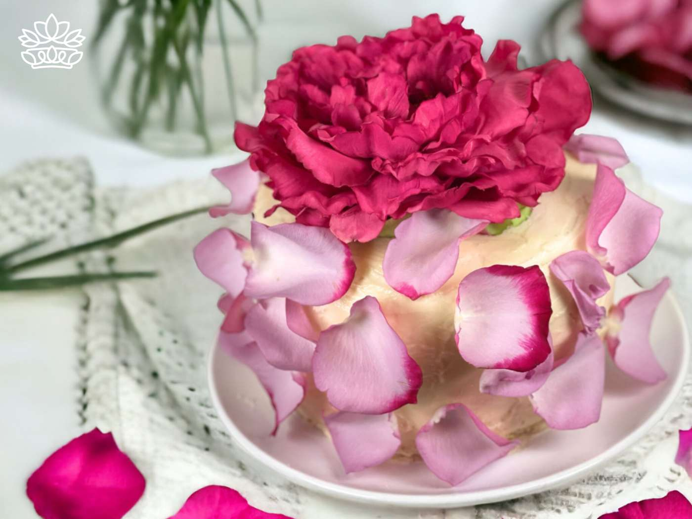 A sumptuous cake crowned with a lush pink peony and surrounded by scattered rose petals, presented on a delicate lace tablecloth, a signature offering from Fabulous Flowers and Gifts.