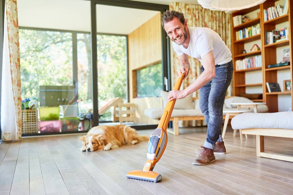 A man is smiling while vacuuming the floor with a relaxed dog lying nearby, suggesting to Use an Air Purifier for Dog