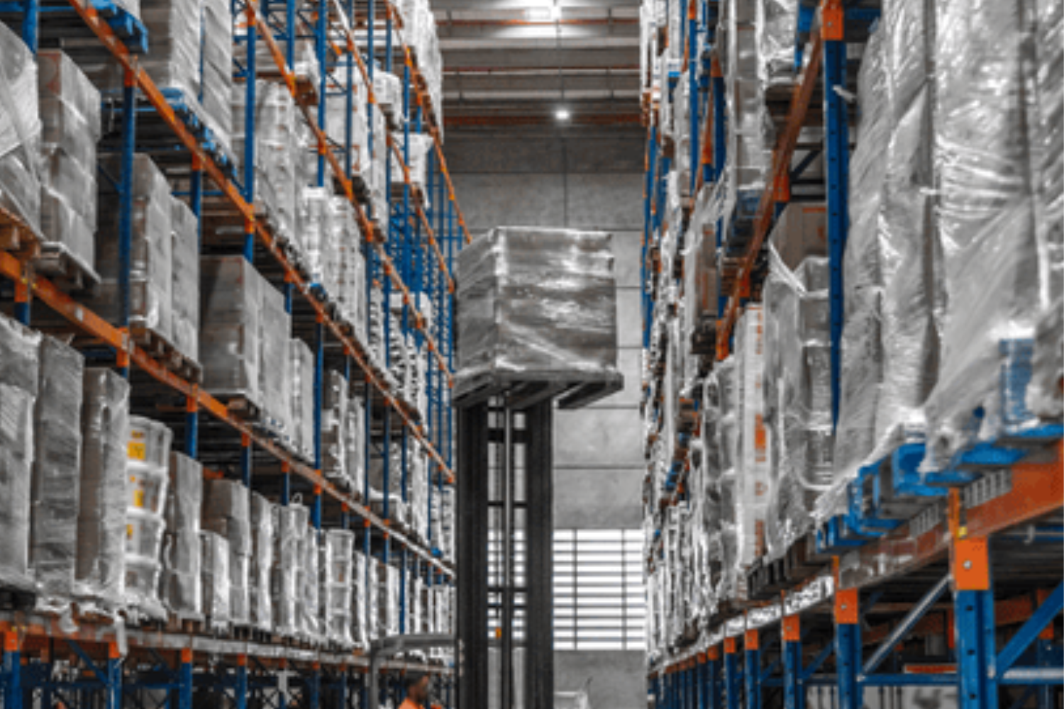 A forklift holds a wrapped pallet in a warehouse with rows of blue and orange shelving filled with wrapped boxes.