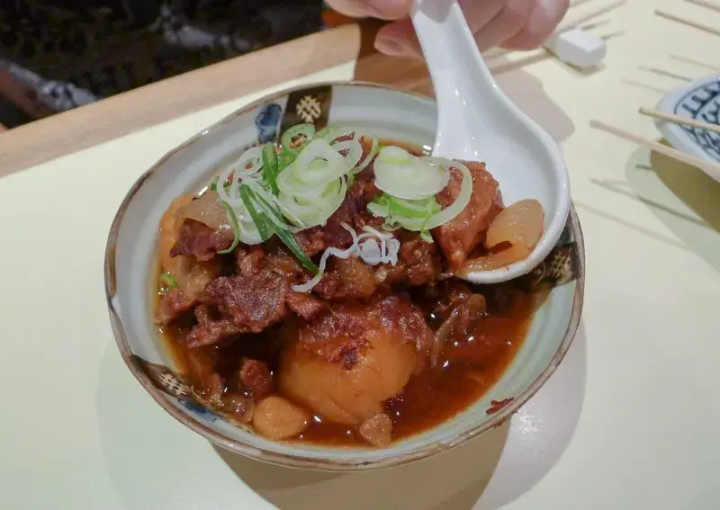 This image shows a small ceramic bowl of Nikujaga, a Japanese beef and potato stew, garnished with sliced green onions and served with a white ceramic spoon. The dish features tender chunks of meat and vegetables simmered in a dark, savory soy-based broth.