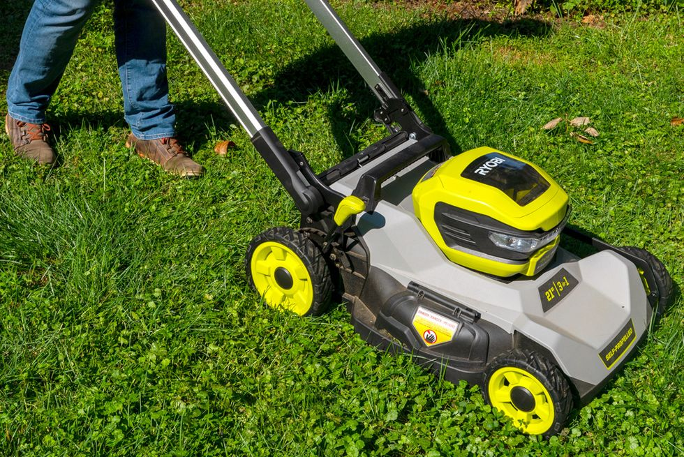 A person is using a Ryobi 21-inch self-propelled lawn mower with a grey and yellow design on a vibrant green lawn. The mower features a sturdy build and is being operated by someone in jeans and brown boots.