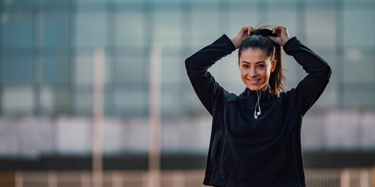 A diet break helps with mindful eating. Woman in black sweater has headphones hanging out of collar while she tightens her ponytail.