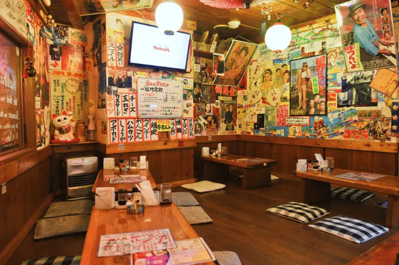 The cozy, retro interior of a Singapore izakaya, featuring low wooden tables with plaid floor cushions for traditional seating, surrounded by walls densely covered in vintage Japanese movie posters, ads, and handwritten menus.