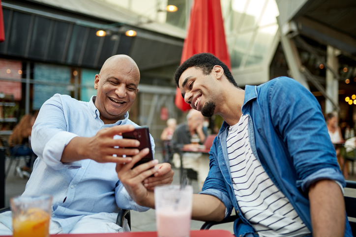 Dad and his adult son smiling over cold drinks and looking at a cell phone. 