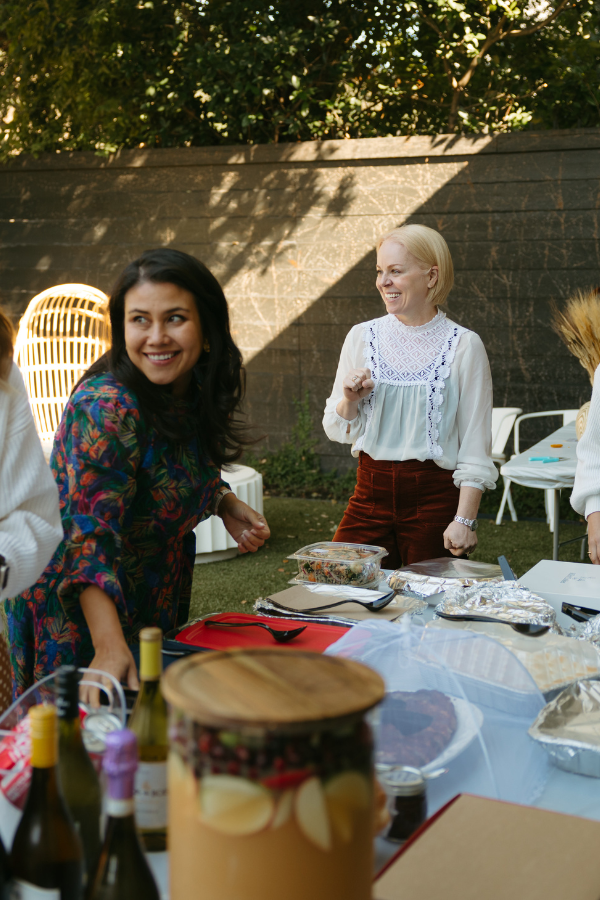 Group of women laughing and serving food at an outdoor gathering, standing around a table with wine bottles, dishes, and shared platters in a sunny backyard.