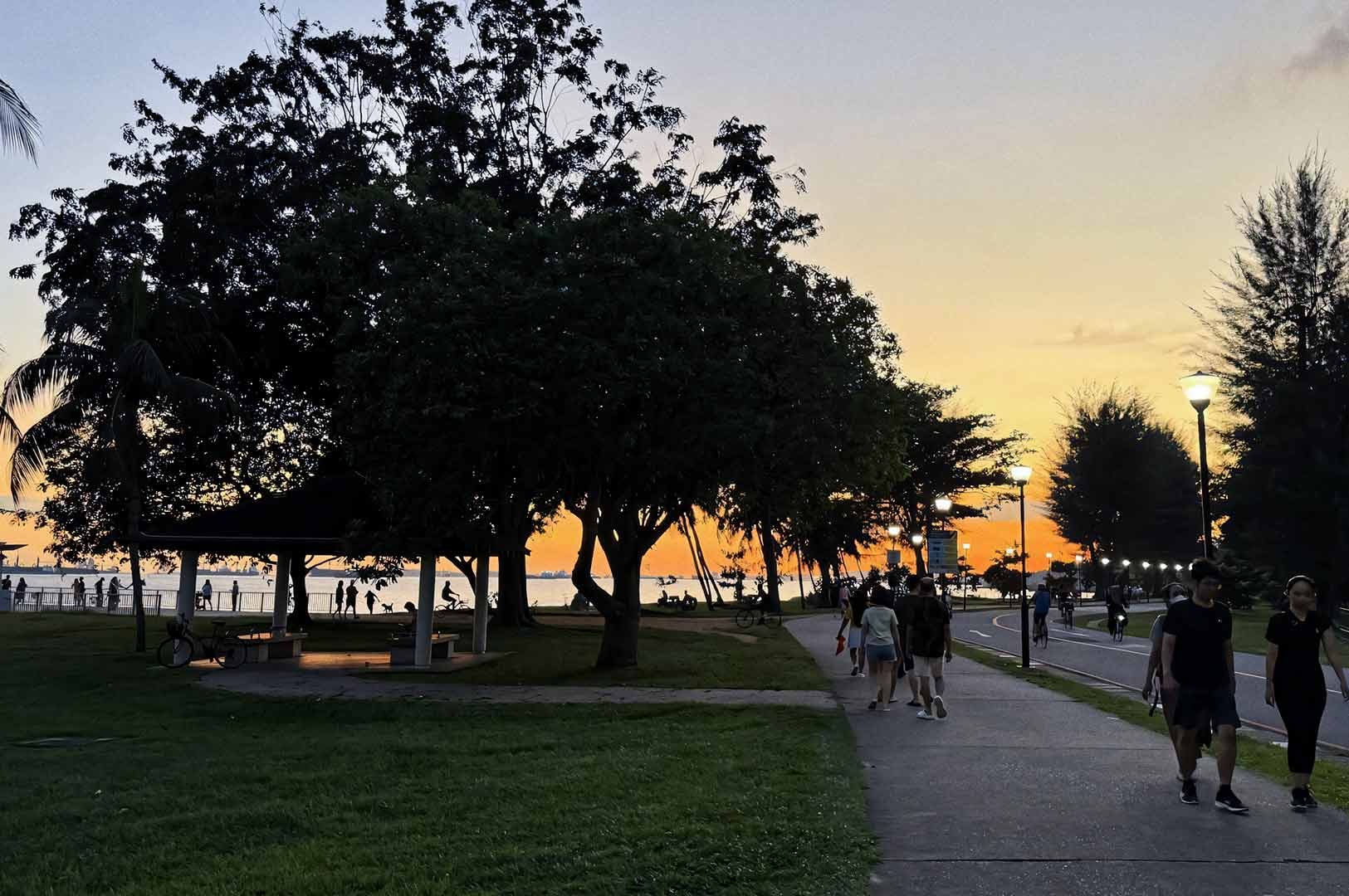 Sunset at East Coast Park in Singapore, with the sky glowing orange and fading into blue. Silhouetted trees line the shore, and people stroll, cycle, and relax along the walking and cycling paths. A pavilion sits near the water as the sun sets over the sea.