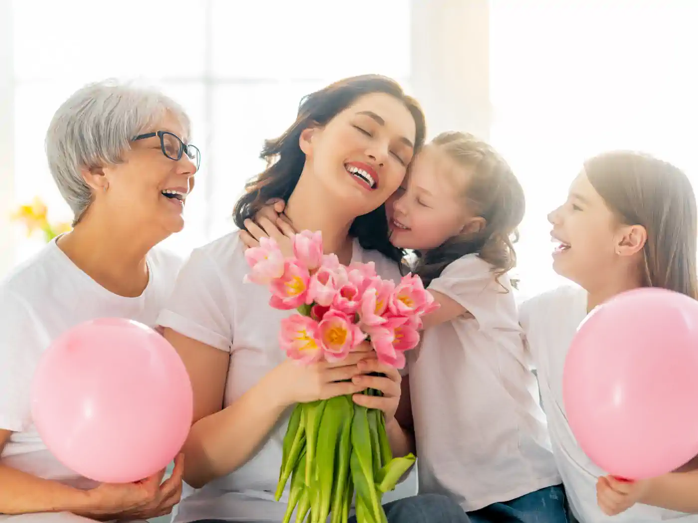 Three generations of women, including a grandmother, mother, and two daughters, hugging and holding flowers and balloons. Fabulous Flowers and Gifts. Collection: Women's Day.