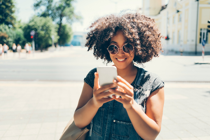 Smiling young woman with dark curly hair and sunglasses sending a text in the sunshine.  