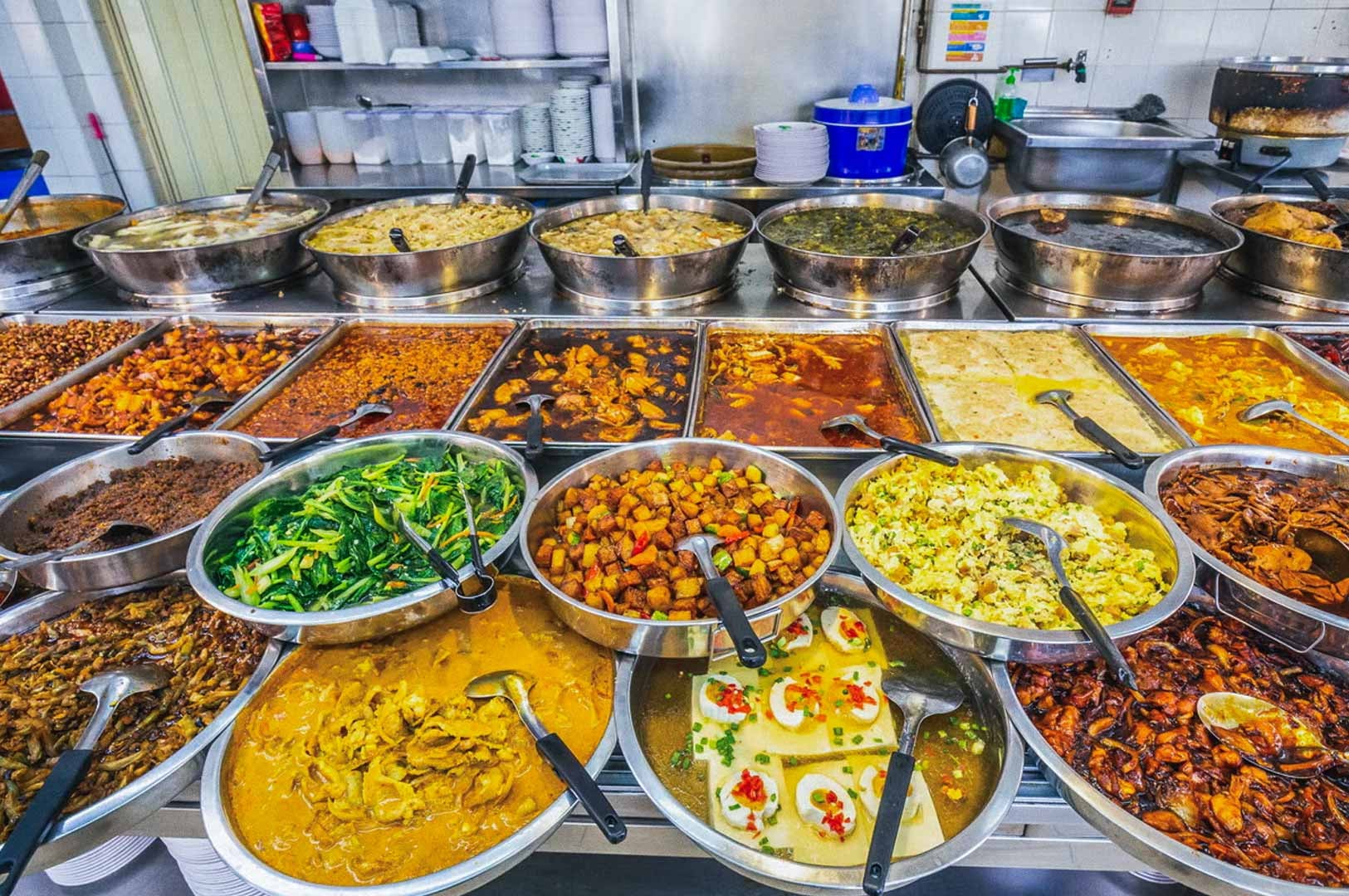 Wide selection of Asian dishes displayed in metal trays at a hawker food stall.