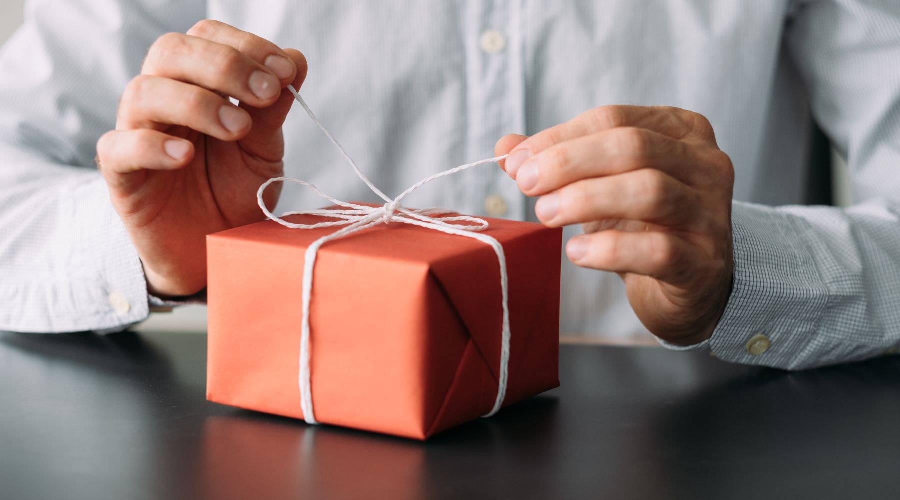 Close-up of a person tying a white string around a small red gift box on a dark desk