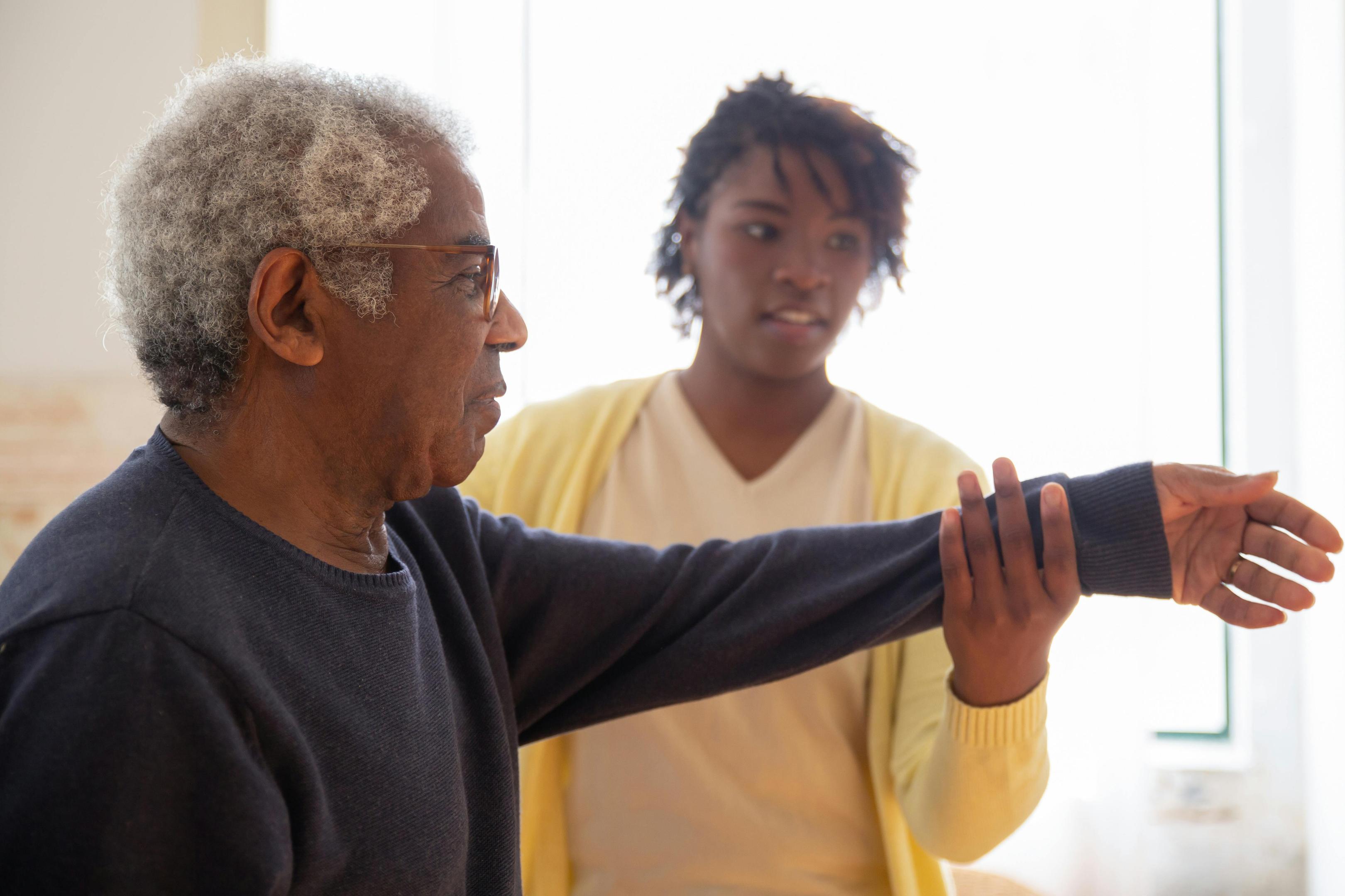 An elderly man with a caregiver holding his hand out in the air to help with his mobility.