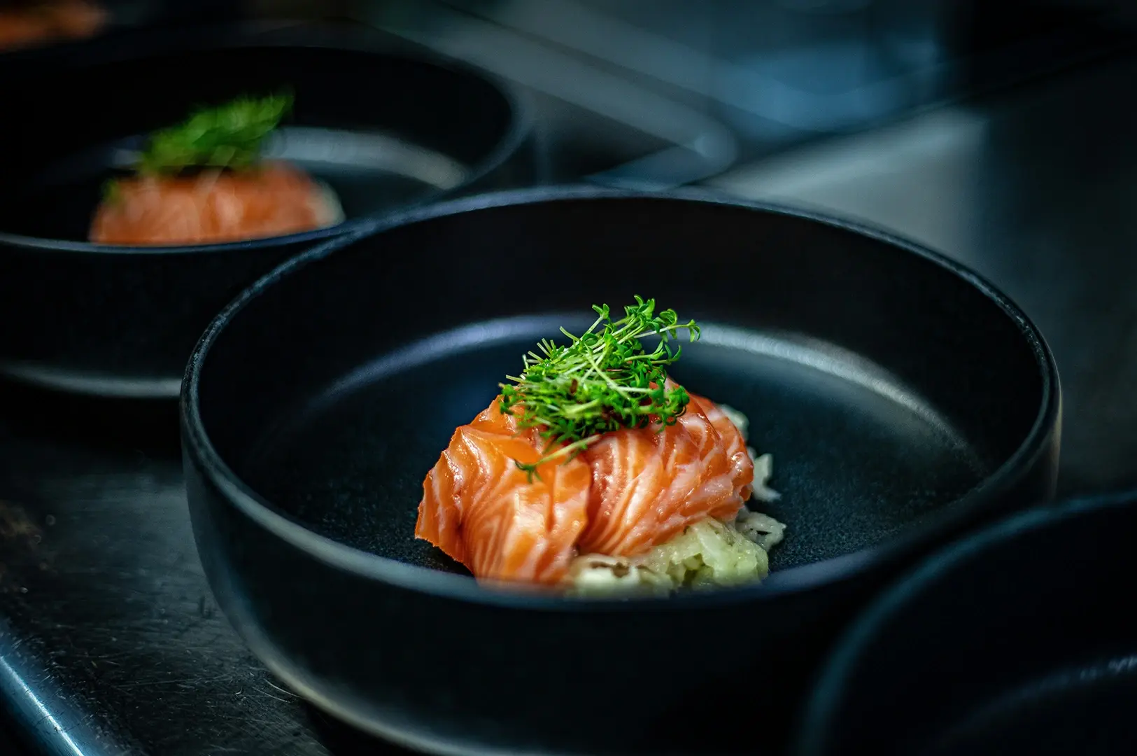 Fresh salmon sashimi garnished with green microgreens and served in a dark ceramic bowl.
