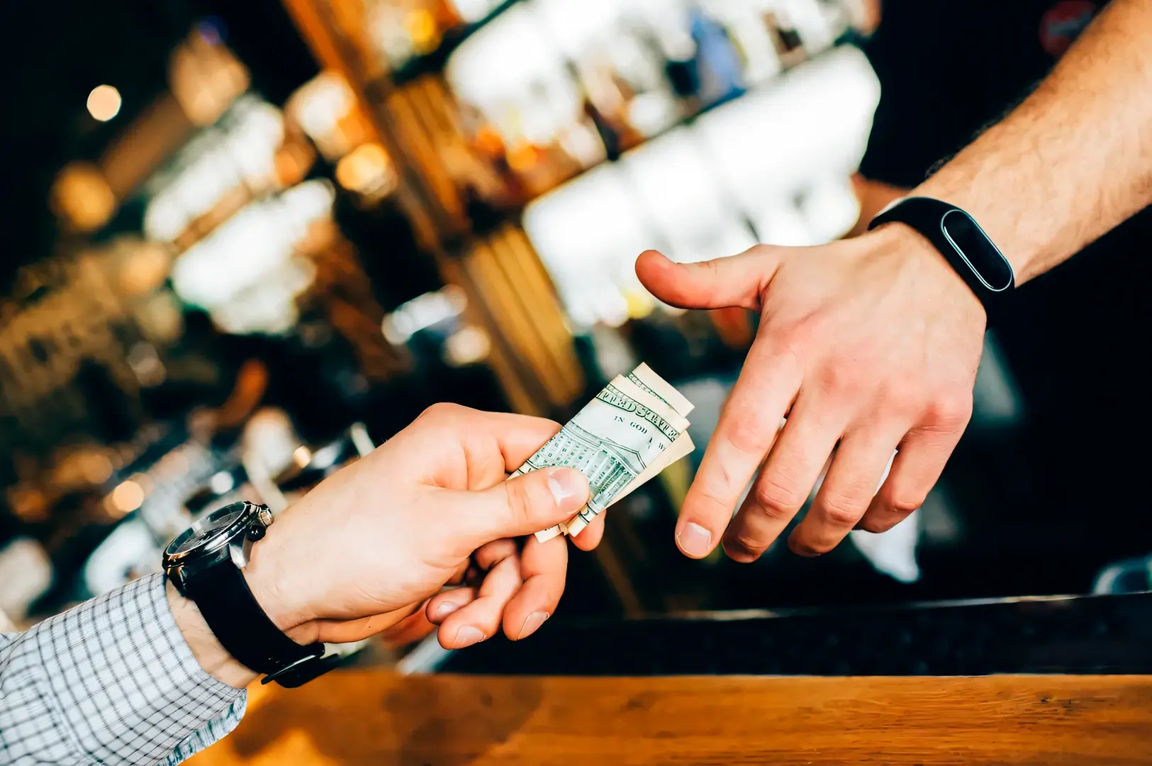 Close-up eye-level shot of one person handing folded cash across a wooden bar counter to another person, with illuminated bar shelves and glassware softly blurred in the background.