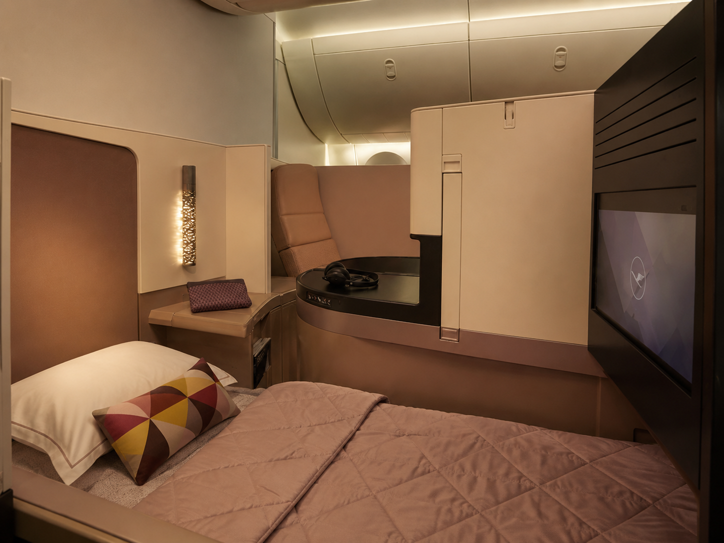 Luxury first class airplane suite with a fully made bed, quilted bedding, decorative pillow, privacy walls, headphones and an entertainment screen.