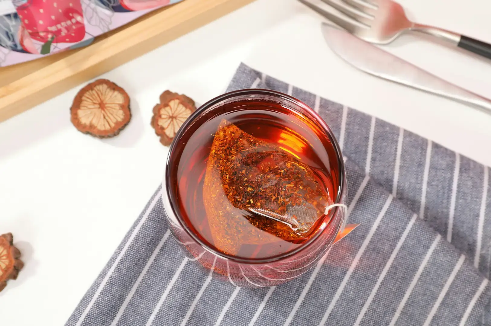 A top-down view shows a clear glass filled with rich reddish-amber tea steeping from a mesh pyramid teabag, resting on a grey and white striped fabric napkin. Dried fruit slices are scattered nearby on the clean white surface, while a wooden tray edge and silver cutlery frame the upper corner of the scene.