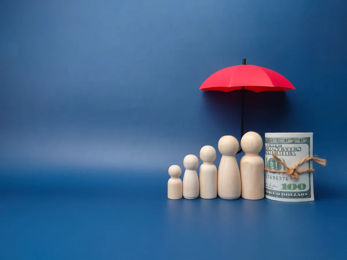 A visual representation of family financial protection with wooden figurines of different sizes arranged under a red umbrella next to a roll of money against a blue background.