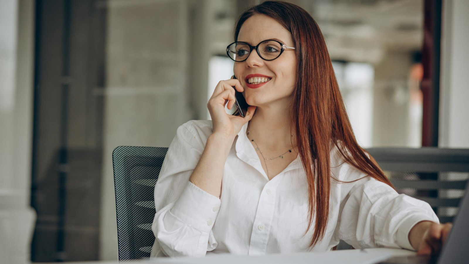 Smiling woman in glasses talking on a smartphone while working at a desk in a modern office.