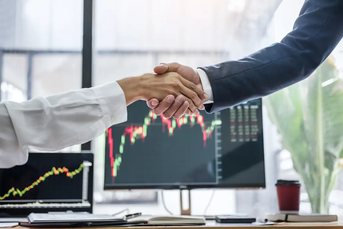 Two people shaking hands in front of financial charts on screen, representing the successful outcome of a meeting initiated with key interview questions for business brokers.
