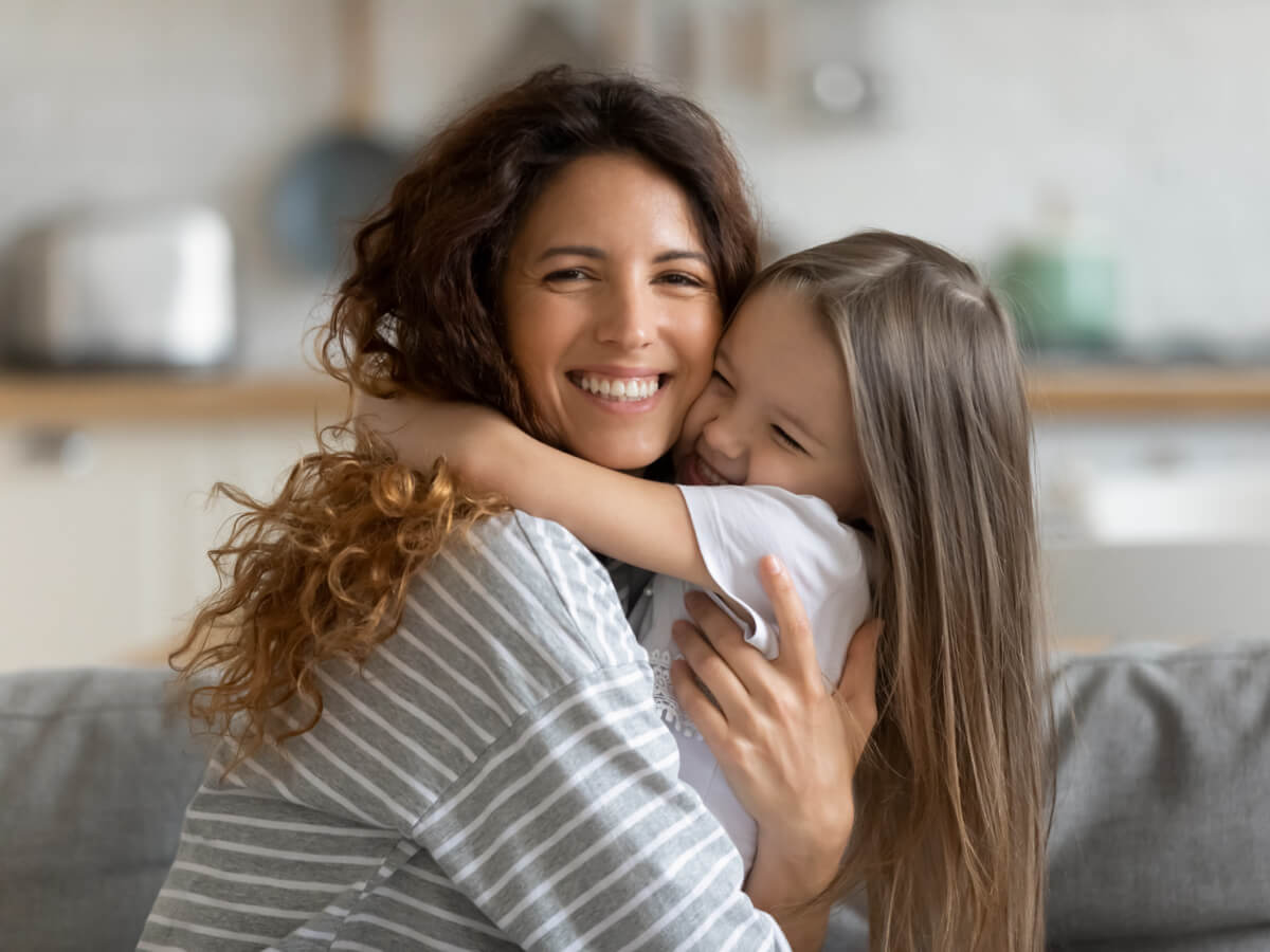 White woman smiling and hugging a little girl, also smiling while sitting on the coach