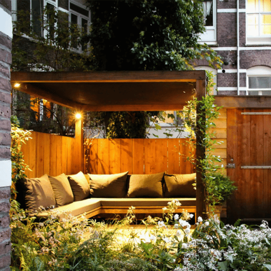 Cozy outdoor seating area with a wooden pergola illuminated by smart LED bulbs, featuring warm overhead lighting and surrounded by lush garden greenery.