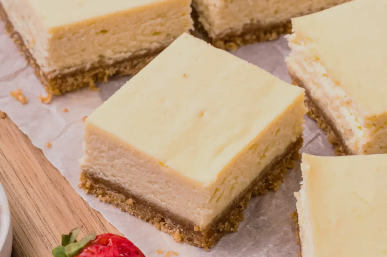 Cheesecake squares with a graham cracker crust, some pieces visible with a strawberry on the side.