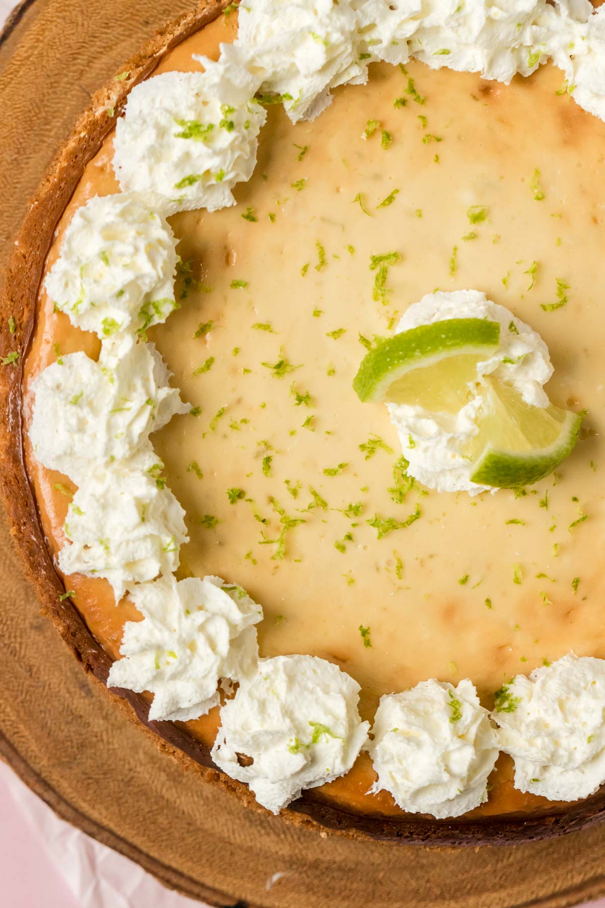 top down view of a whole key lime cheesecake topped with whipped cream and key lime zest and lime slices on a wood platter 